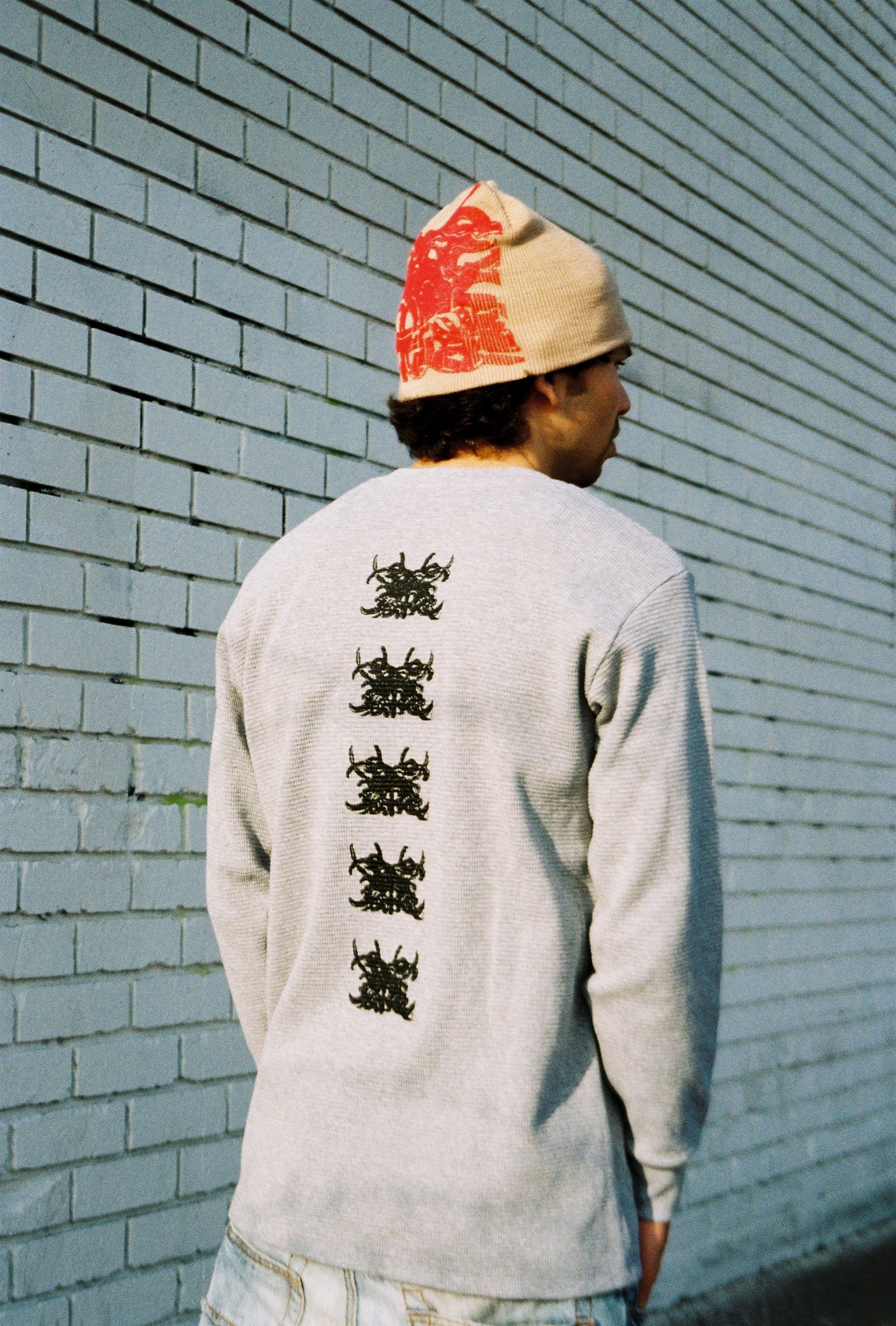 Person wearing a light gray long-sleeve shirt with black designs on the back, standing against a light blue brick wall.