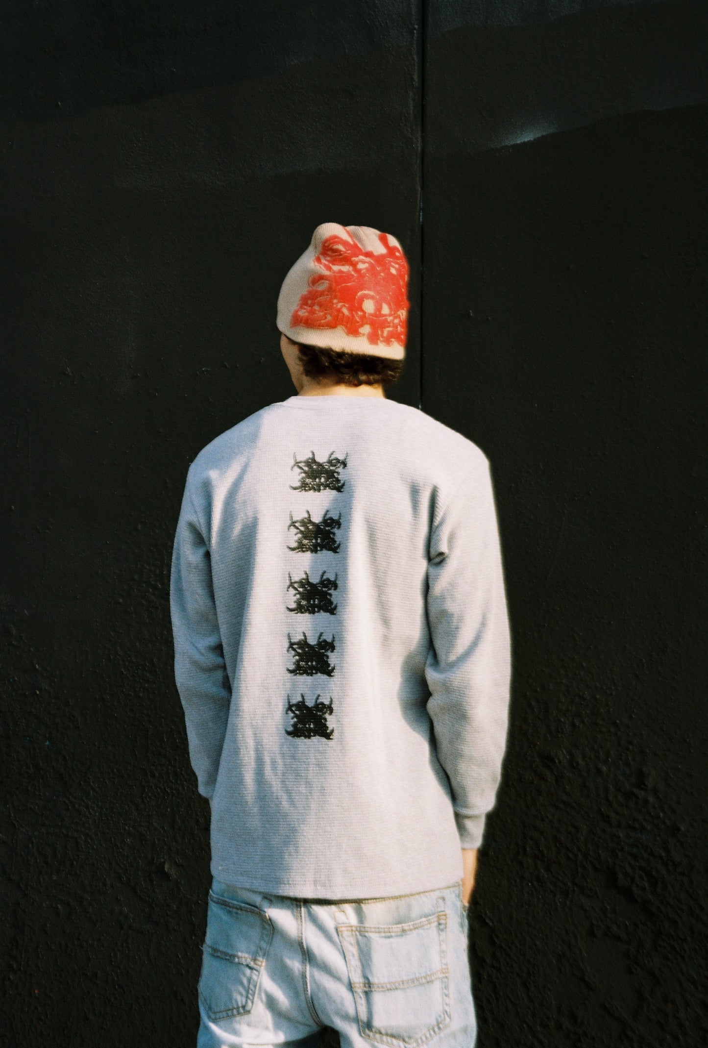 Person wearing a light gray long-sleeve shirt with black graphics on the back, standing against a dark wall.